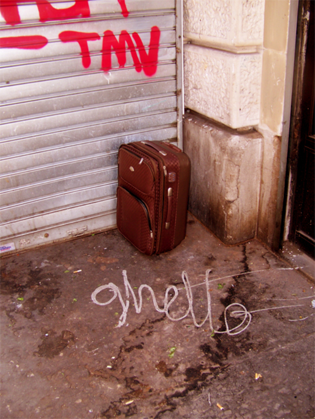 Ein brauner Koffer steht an einer Ecke neben einer Rollladen-Tür in einer unattraktiven Umgebung.