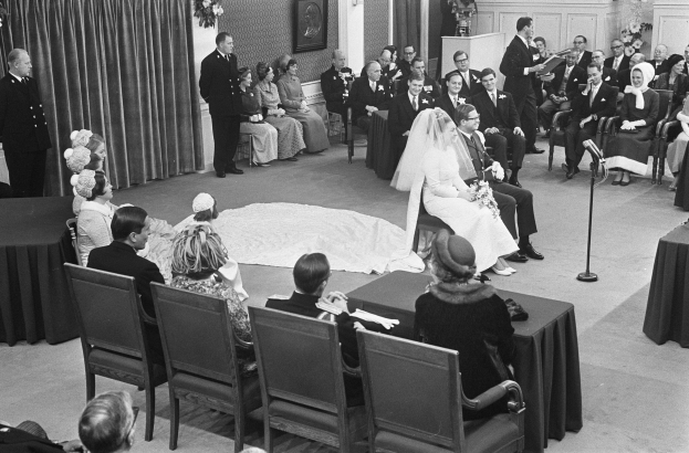 Schwarzes und weißes Foto einer Hochzeitszeremonie mit Menschen, die sitzen und stehen, einem Mann mit Mikrofon, einer Frau mit Blumenstrauß, einem Podium mit Mikrofon, Blumenschmuck, einem Vorhang, einem Foto auf der Wand und einer Tür im Hintergrund.