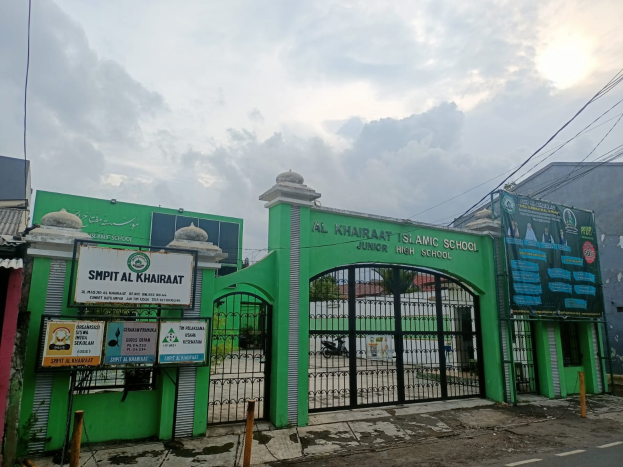 Der Eingang der Al Khairat Islamischen Schule, mit einem gr├╝nen Tor und einem Schild darauf, sowie Pfosten und Bretter, mit Geb├Ąuden, B├Ąumen und einem klaren blauen Himmel im Hintergrund.