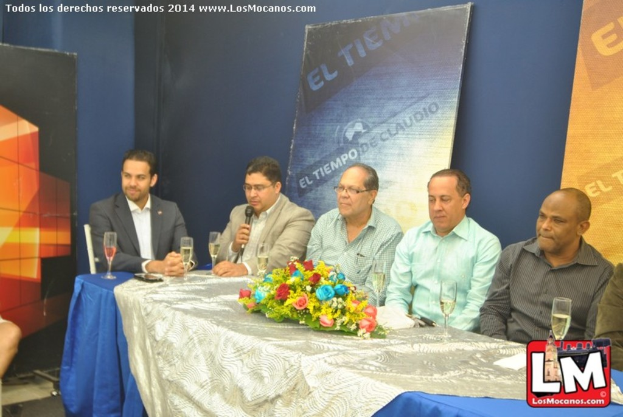 Sechs Personen sitzen um einen Tisch mit Blumenstrauß und Gläsern, ein Mann in schwarzem Anzug und weißem Hemd links, mit Bannern und Plakaten im Hintergrund.