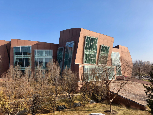 Großes modernes Gebäude mit vielen Fenstern, umgeben von Bäumen, Bänken, Gras und einem Laternenmast, mit anderen Gebäuden und einem klaren blauen Himmel im Hintergrund; als Campus der University of Illinois at Chicago identifiziert.