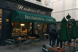 Eine Gruppe von Menschen vor einem Cafe mit einer Tafel, auf der 'Ruskin's Cafe' steht.