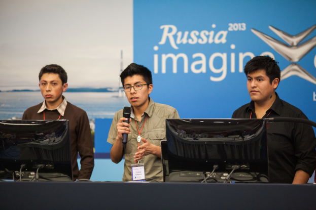 Drei Jungs stehen neben einem Schreibtisch mit zwei Computern, einer spricht in ein Mikrofon und tr├Ągt ID-Karten mit roten Markierungen, vor einer Wand mit einer Werbetafel, auf der "Russia Imagine 2013" steht.