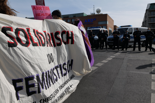 Eine Gruppe von Menschen marschiert auf einer Straße, hält eine Fahne mit der Aufschrift "Solidarität und Feminismus" und fährt vorbei an parkenden Fahrzeugen und Gebäuden unter einem klaren blauen Himmel.