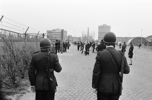 Zwei Polizisten in Helmen und mit Waffen in der Hand stehen nebeneinander auf einem Bürgersteig, links davon ein Zaun und Pflanzen, im Hintergrund Gebäude, Pfähle und einen klaren Himmel.