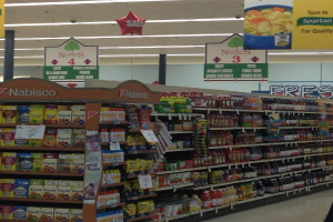 Ein Supermarkt-Innenraum mit deckenmontierten Leuchten und Beschriftungen, der Lebensmittel auf Regalen zeigt.