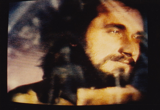 Ein Mann mit langem dunklem Bart und Haar, der ein weißes Hemd trägt, erscheint auf einem Fernseher mit ernstem Gesichtsausdruck und direktem Blick.