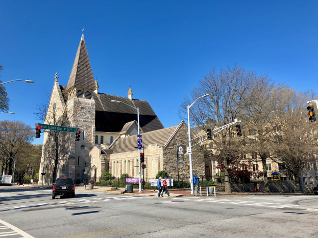 St. Lukes Episkopalkirche, eine große Kirche mit einem Turm, steht an einer Straßenecke umgeben von Gebäuden, Fahrzeugen, Fußgängern und Grün unter einem klaren blauen Himmel.