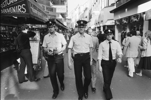 Drei Polizeibeamte in Uniform gehen eine belebte Straße mit hohen Gebäuden, Geschäften und Passanten entlang.