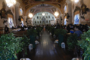 Eine Hochzeitszeremonie in einer Kirche mit Gästen auf Bänken sitzend und im Raum stehend, Topfpflanzen schmücken den Gang, Skulpturen, Vorhänge an den Fenstern und Kronleuchter an den Wänden, sowie ein Bildschirm im Hintergrund.