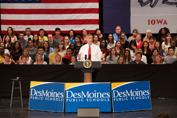 Ein Mann in weißem Hemd und roter Krawatte steht an einem Rednerpult mit drei Tafeln, einem Hocker mit Mikrofon und einer sitzenden Menge im Hintergrund, mit einer Flagge und zusätzlichem Text.