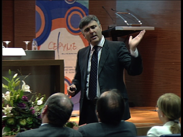 Ein Mann in einem schwarzen Blazer steht an einem Podium mit Mikrofonen und spricht in eines daneben, neben einem Blumenarrangement und einem Tisch mit einem Buch und einer Wasserflasche; zwei Männer und eine Frau sitzen in Stühlen davor, mit einer braunen Wand und einer bunten Tafel mit Text im Hintergrund.