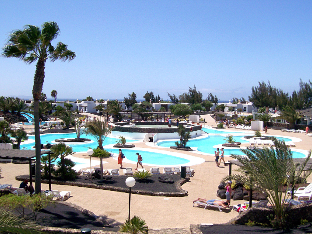 Ein Resort mit Pools umgeben von Bäumen, Menschen, die dazwischen gehen, Gebäude im Hintergrund und ein klarer Himmel darürber.