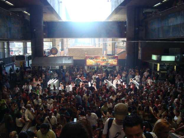 Eine Menschenmenge vor Marktständen mit fahrenden Fahrzeugen im Hintergrund und Säulen an den Wänden.