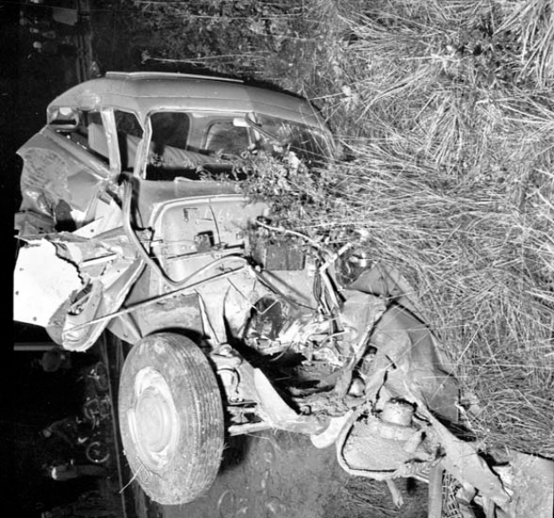 Ein Schwarz-Weiß-Foto eines schwer beschädigten Autos am Rande einer Straße, mit einer zerbeulten Front, zersplitterten Fenstern, einem Baum im Hintergrund und verstreuten Gegenständen um die Szenerie.