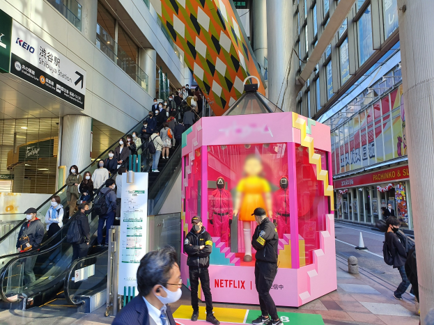 Eine Gruppe von Menschen steht vor einem Gebäude mit Glaswänden und Texttafeln, einige tragen Masken, während eine Rolltreppe mit Fahrgästen zu sehen ist. In der Mitte steht ein pinker Kasten mit der Aufschrift "Netflix's new Netflix booth in Tokyo".