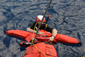 Retter in rotem Lebensretter und Helm im Wasser mit einem Sicherheitsseil am Rücken.