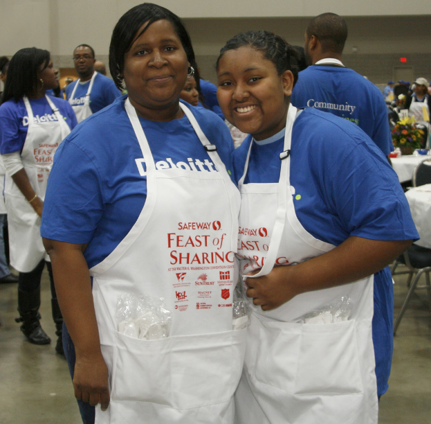 Zwei Frauen in Kochschürzen mit Gegenständen in den Taschen stehen in einem Raum mit Menschen, Tischen, Stühlen, einer Blumenvase und Schüsseln, unter roter Beleuchtung an der Wand.
