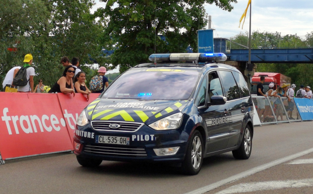 Polizeiauto fährt an einer Menge vorbei, die sich an einem Geländer mit Bannern befindet, daneben Bäume, eine Brücke, eine Fahne und einen bewölkten Himmel im Hintergrund.