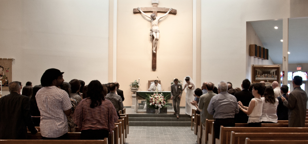 Eine kleine Gruppe von Menschen nimmt an einer Hochzeit teil, im Hintergrund ist eine Statue von Jesus Christus an einer Wand befestigt.