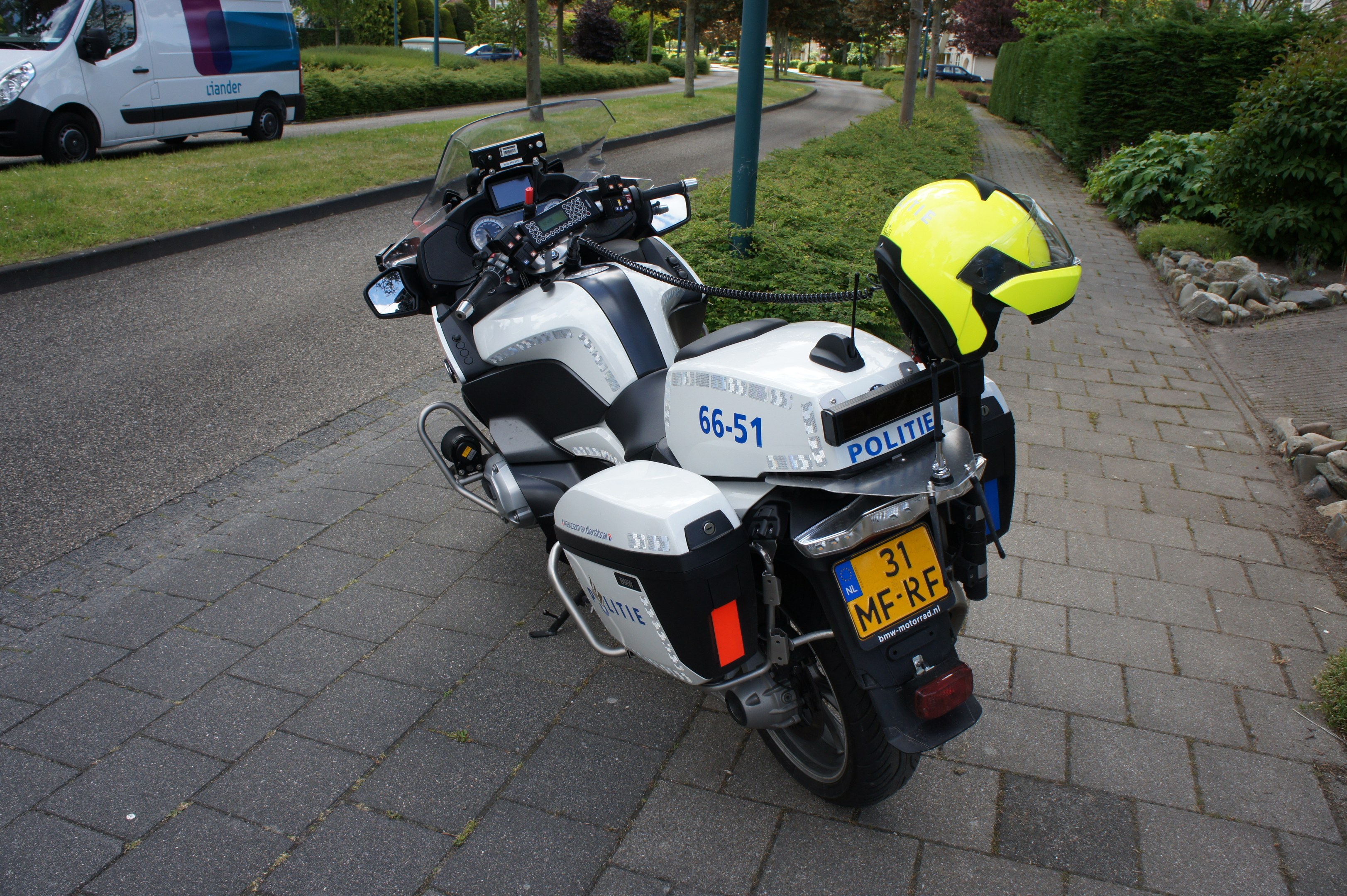 Ein polizeiliches Motorrad am Straßenrand geparkt mit einem Helm darauf, umgeben von Bäumen, Pfählen, Gebäuden, Pflanzen, Gras, Steinen und anderen Fahrzeugen.
