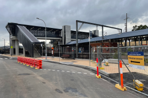 Ein Bahnhof mit einer Brücke, Straßenlaternen, Strommasten, Stromkabeln, Bäumen, Gebäuden, Fahrzeugen, Barrierepfosten, Schildern, Bänken und einer bewölkten Himmel