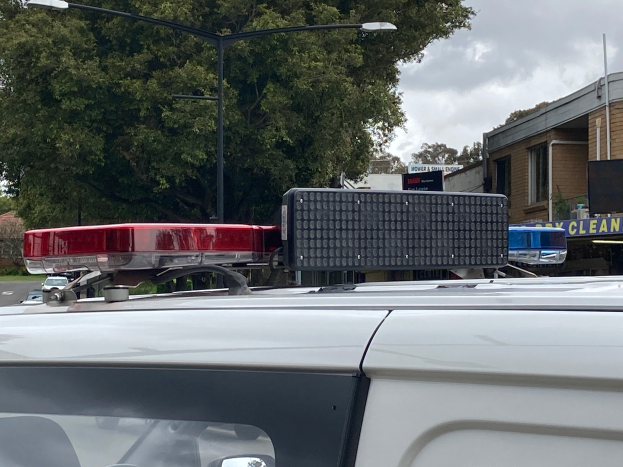 Ein Polizeiwagen mit eingeschaltetem Licht steht auf der Seite einer Straße umgeben von Gebäuden, Bäumen, Polen und Texttafeln, mit einem bewölkten Himmel im Hintergrund, wahrscheinlich nach einem tödlichen Schulschießen in Brisbane.
