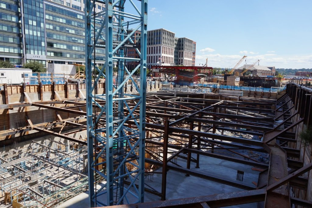 Das Bild zeigt eine Baustelle mit Gebäuden und Büros links, Eisenstangen und Maschinen rechts und diverser Ausrüstung und Kränen in der Mitte, unter einem Himmel im rechten oberen Eck.