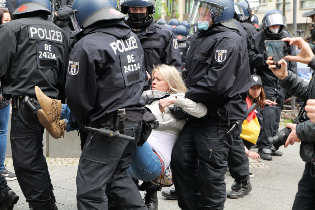 Eine Gruppe von Polizisten in Einsatzausrüstung umgibt eine Frau in einem weißen und blauen Kleid, während sie sie eskortieren und eine Person rechts ein Foto macht, mit Gebäuden, Bäumen und anderen Menschen im Hintergrund.