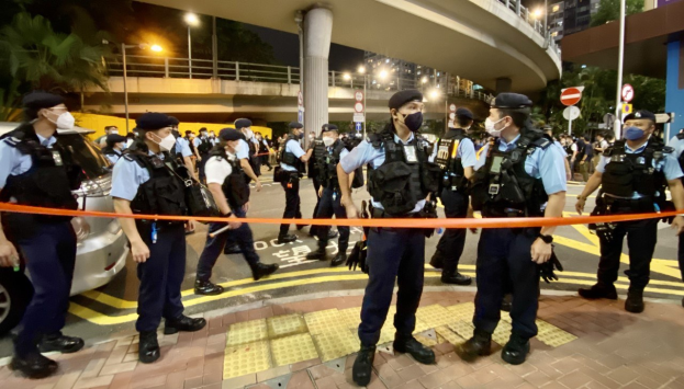 Polizisten in Uniform und Masken vor einer Menge mit Fahrzeugen und Gebäuden im Hintergrund, wahrscheinlich nach einer Demonstration in Hong Kong.