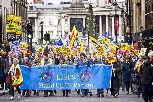 Eine Gruppe von Menschen marschiert mit einer 'Stand Up to Racism'-Schleife die Straße entlang, mit Gebäuden, Bäumen, Laternen, Verkehrszeichen und einer Statue im Hintergrund bei bewölktem Himmel.