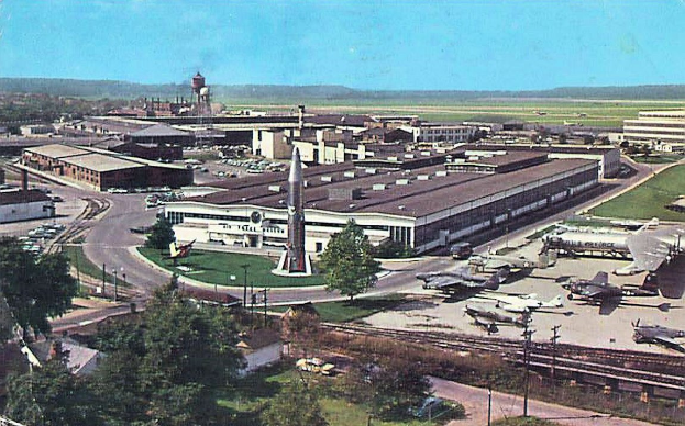 Altes Schwarz-Weiß-Foto eines Flughafens mit geparkten Flugzeugen, Gebäuden, Bäumen, Straßen, Fahrzeugen, Strommasten, Gras, Bahnschienen, Türmen, fernen Hügeln und einem klaren blauen Himmel, mit Text unten.