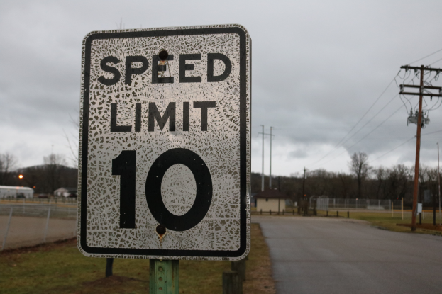 Ein Geschwindigkeitsbegrenzungsschild am Straßenrand, umgeben von Gras, Strommasten mit Drähten, einem Zaun, Häusern, Bäumen und einem Himmel mit Wolken im Hintergrund.