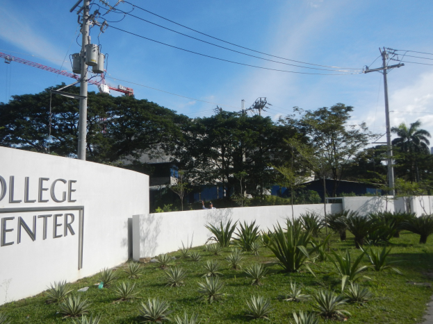 Eingang zu einem College-Zentrum, markiert von einem "College-Zentrum"-Schild, umgeben von Grün, einem Weg, Strommasten, Bäumen, Gebäuden, einem Kran und einem bewölkten Himmel.