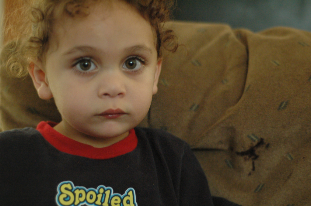 Ein Kind mit lockigen Haaren, das ein schwarzes Hemd mit dem gelben Wort "Spoiled" trägt, vor einem braunen Stoffhintergrund.