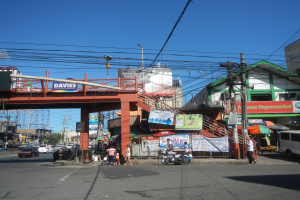Eine belebte Stadtstraße mit einer Brücke darüber, Fahrzeuge und Fußgänger auf der Straße, Strommasten mit Drähten, Gebäude mit Fenstern, Banner, Laternenpfähle, Bäume und einen klaren blauen Himmel.