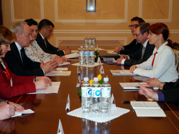 Neun Personen sitzen um einen Tisch in einem Konferenzsaal, mit Flaschen, Tüchern, Büchern und Stiften auf dem Tisch, eine Person hält einen Stift, und ein gerahmtes Bild im Hintergrund.