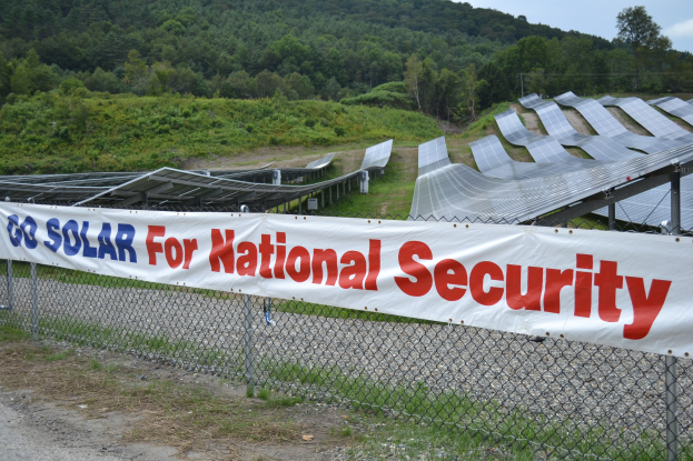 Ein Zaun mit einer "Gehen Sie Solar für nationale Sicherheit"-Aufschrift vor einem Solarpanel-Feld, Bäumen, Pflanzen, Gras und einem klaren blauen Himmel.