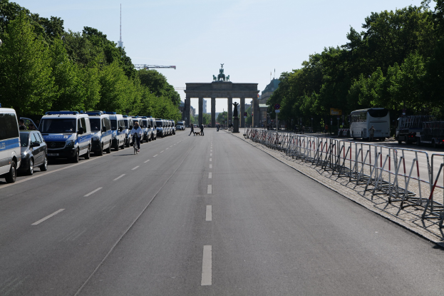 Lange Reihe von Polizeiwagen auf der Seite einer Straße vor dem Brandenburger Tor in Berlin, Deutschland geparkt, mit Menschen auf Fahrrädern und auf der Straße stehend, Barrieren, Bäume, ein Bogen mit Statuen im Hintergrund und sichtbarer Himmel.