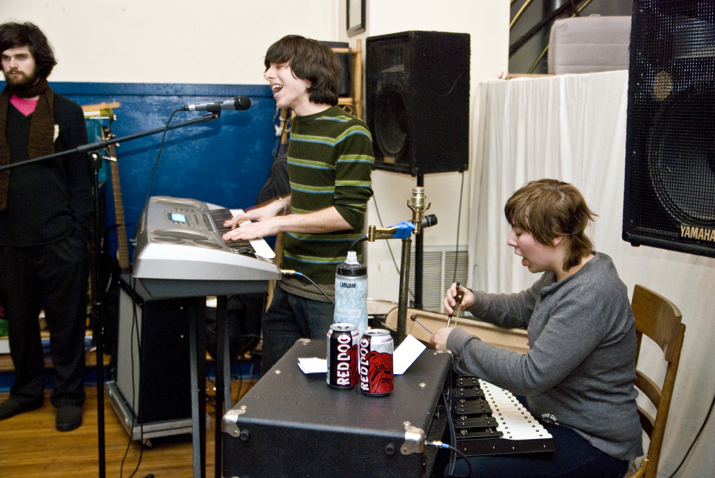 Drei Männer, einer steht links, ein anderer spielt Piano und singt, und der dritte singt und hält Getränkedosen auf einem Tisch, mit einem Lautsprecher und einem Vorhang im Hintergrund.