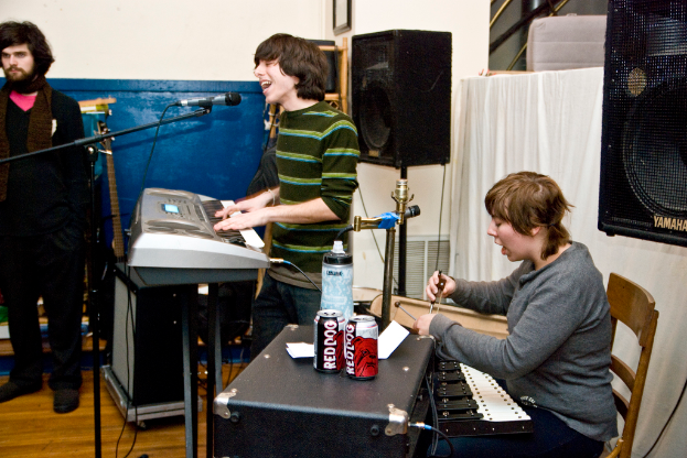 Drei Männer, einer steht links, ein anderer spielt Piano und singt, und der dritte singt und hält Getränkedosen auf einem Tisch, mit einem Lautsprecher und einem Vorhang im Hintergrund.