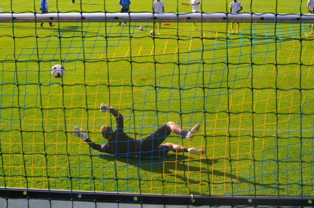 Ein Fussballspieler hält den Ball vor einem Tor, mit einem Netz im Vordergrund und einigen Menschen im Hintergrund sowie mehreren Bällen auf dem Boden.