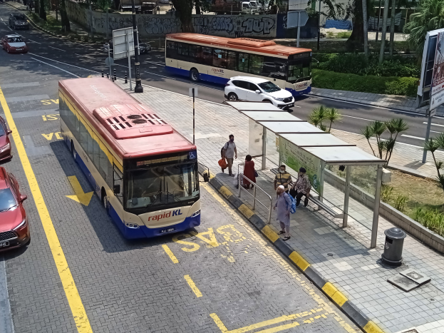 Stadtstraße mit fahrenden Autos, Bussen, Bushaltestelle, Fußgängern auf dem Gehweg, Strommasten, Schilder, Bäume, Gras und umliegende Gebäude.