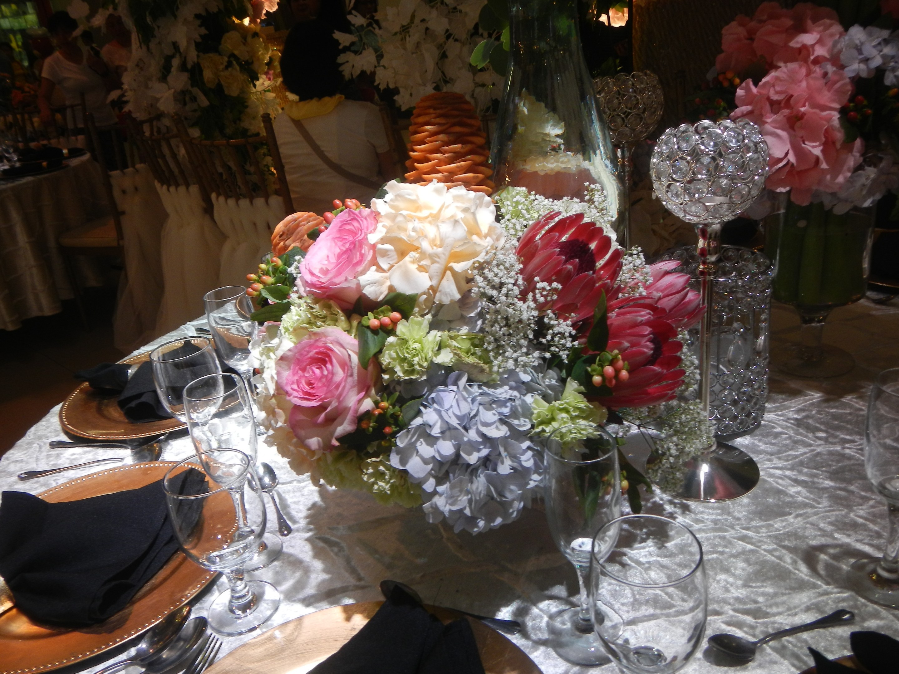 Ein festlich gedeckter Esstisch mit Tellern, Besteck, Gläsern, Servietten und Blumenvasen, mit einem Blumenarrangement in der Mitte, und Stühlen und Tischen im Hintergrund.