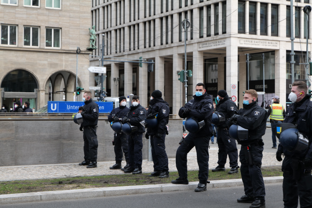 Eine Gruppe von Polizisten in schwarzen Uniformen und Masken steht vor einem Berliner Gebäude mit Glasfenstern, Säulen und Gras, einige halten Helme, während Laternenpfähle, Verkehrsampeln, Schilder mit Text, eine Statue und andere Menschen im Hintergrund zu sehen sind.