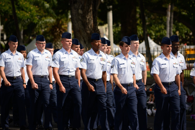 Männer in Uniformen gehen auf einer Straße mit einem Eisenzaun dahinter, Bäume und Pfosten im Hintergrund.