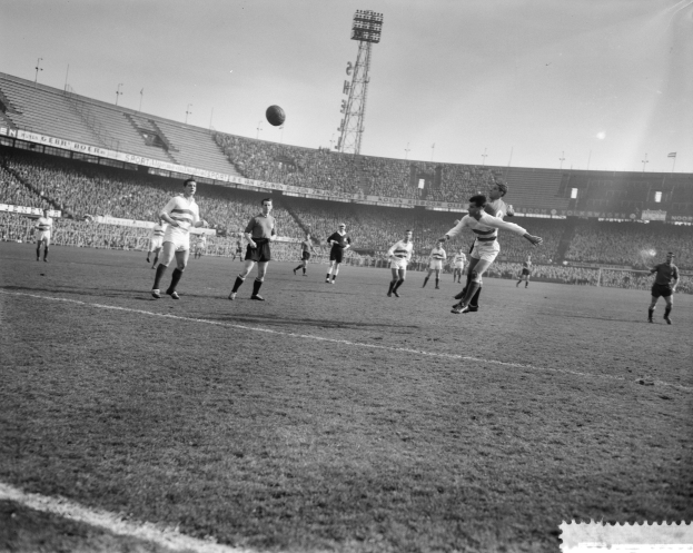 Eine Gruppe Männer spielt Fußball auf einem Feld mit Stadion, Flutlicht und einem klaren Himmel im Hintergrund.
