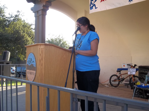 Eine Frau in einem blauen T-Shirt steht vor einem Mikrofon, mit Bäumen, Himmel, Autos, einer Fahne und einem kleinen Fahrrad im Hintergrund.