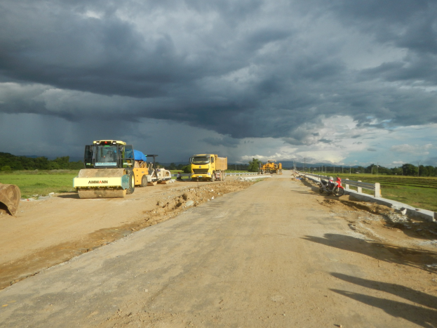 Straßenbaustelle mit Fahrzeugen, einem Zaun auf der rechten Seite, Gras auf dem Boden, Bäumen im Hintergrund und einem bewölkten Himmel.