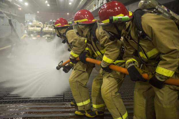 Feuerwehrleute in Helmen und Handschuhen sprühen Wasser auf ein Feuerwehrauto, während andere Ausrüstung und Beschriftungen im Hintergrund zu sehen sind.